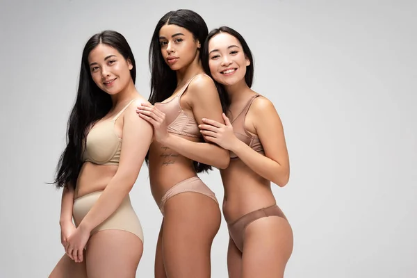 Tres hermosas chicas multiétnicas en lencería mirando a la cámara aislada en gris, concepto de positividad corporal - foto de stock