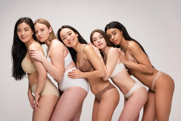 Cinco jóvenes y felices mujeres multiétnicas en ropa interior apoyadas una en la otra aisladas en gris, concepto de positividad corporal - foto de stock