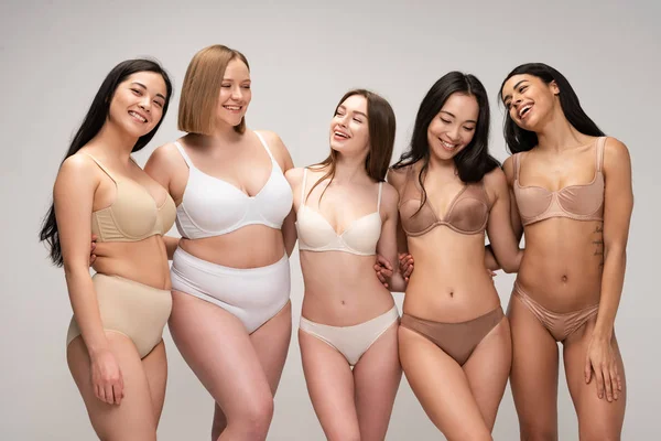 Пять улыбающихся мультикультурных девушек в женском белье обнимаются, позируя перед камерой, изолированной на сером, концепция позитивности тела — стоковое фото