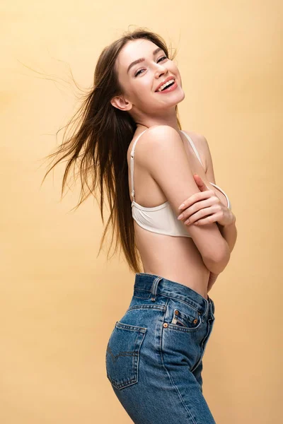 Щаслива молода жінка в блакитних джинсах і бюстгальтер посміхається і дивиться на камеру ізольовано на бежевому — стокове фото