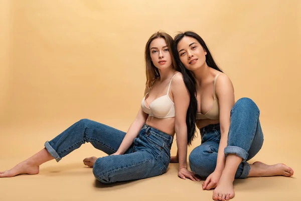 Chicas bonitas felices en pantalones vaqueros azules y sujetadores sentados y mirando a la cámara, concepto de positividad corporal - foto de stock