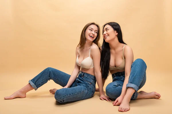 Mujeres jóvenes felices sentados en pantalones vaqueros azules y sujetadores y mirándose entre sí, concepto de positividad corporal - foto de stock