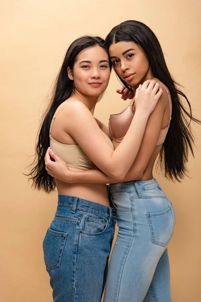 Deux filles multiculturelles heureuses en jeans bleus et soutien-gorge étreignant et regardant la caméra isolée sur beige, concept de positivité du corps — Photo de stock