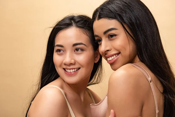 Dos chicas muy multiculturales sonriendo y mirando hacia otro lado aislado en beige - foto de stock