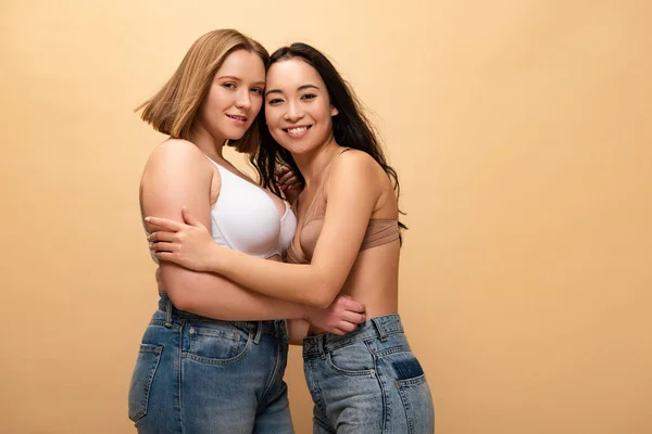 Glücklich übergewichtige Mädchen und schlanke asiatische Frau, die sich umarmt, während sie in die Kamera schaut, isoliert auf beige, Body Positivity Konzept — Stockfoto