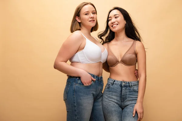 Glücklich übergewichtige Mädchen und schlanke asiatische Frau umarmen, während sie in die Kamera isoliert auf beige, Body Positivity Konzept — Stockfoto