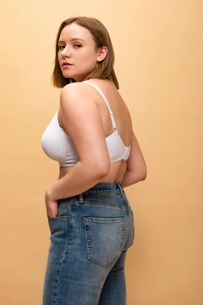 Pretty overweight girl in blue jeans and bra looking at camera isolated on beige, body positivity concept — Stock Photo
