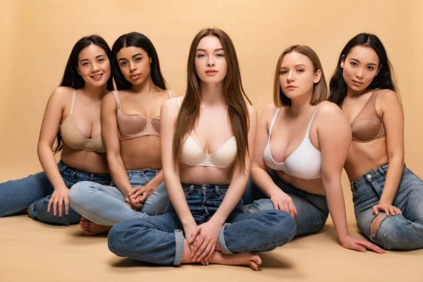 Five beautiful multicultural girls in blue jeans and bras sitting and looking at camera, body positivity concept — Stock Photo