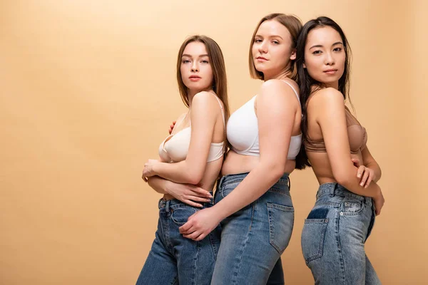 Tres mujeres bastante multiculturales en pantalones vaqueros azules y sujetadores posando en la cámara aislada en beige, concepto de positividad corporal - foto de stock