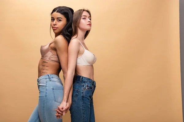 Dos chicas muy multiculturales de pie espalda con espalda y cogidas de la mano aisladas en beige - foto de stock