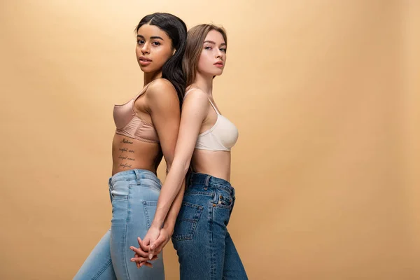 Zwei schöne multikulturelle Frauen, die Rücken an Rücken stehen und die Hände isoliert auf beige halten — Stockfoto