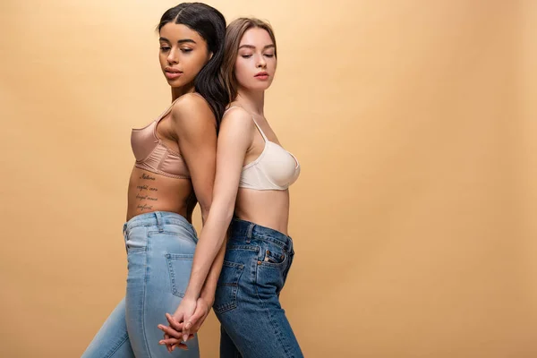 Dos chicas pensativas multiculturales cogidas de la mano mientras se quedan de espaldas aisladas en beige - foto de stock