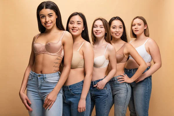 Selective focus of five beautiful multicultural girls in blue jeans and bras smiling and looking at camera isolated on beige, body positivity concept — Stock Photo