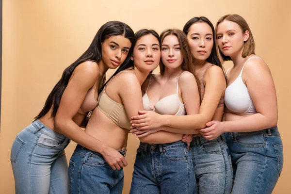 Cinco hermosas chicas multiculturales abrazando y mirando a la cámara aislada en beige, concepto de positividad corporal - foto de stock
