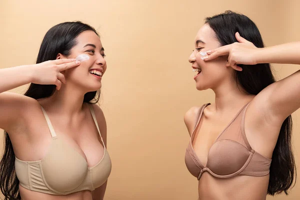 Due ragazze multiculturali felici che applicano crema cosmetica sul viso e si guardano isolate sul beige — Foto stock