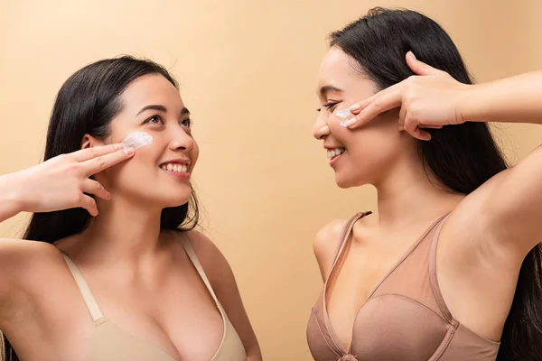 Duas meninas multiculturais alegres aplicando creme cosmético no rosto e sorrindo isolado no bege — Fotografia de Stock