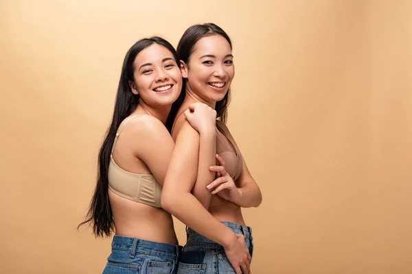 Две улыбающиеся мультикультурные молодые женщины обнимаются, позируя перед камерой, изолированной от бежевого цвета, концепция позитивности тела — стоковое фото