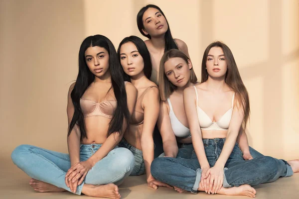 Five multicultural girls in blue jeans and bras sitting in sunlight and looking at camera — Stock Photo