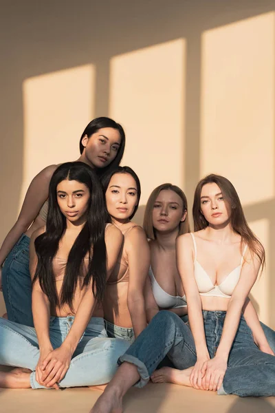 Fünf nachdenkliche multikulturelle Mädchen, die im Sonnenlicht sitzen und in die Kamera schauen — Stockfoto