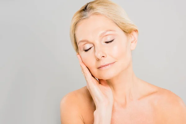 Attractive and blonde mature woman with closed eyes touching her face isolated on grey — Stock Photo