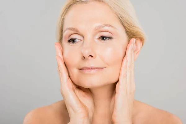 Attraktive und blonde reife Frau, die ihr Gesicht berührt und vor grauem Hintergrund wegsieht — Stockfoto