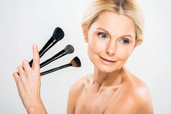 Beautiful and blonde mature woman looking away and holding cosmetic brushes on grey background — Stock Photo