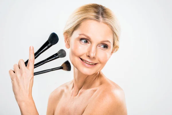 Beautiful and smiling mature woman looking away and holding cosmetic brushes on grey background — Stock Photo