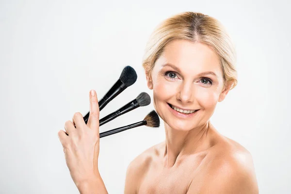 Hermosa y rubia mujer madura mirando a la cámara y la celebración de cepillos cosméticos sobre fondo gris - foto de stock