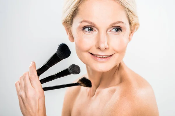 Beautiful and blonde mature woman looking away and holding cosmetic brushes on grey background — Stock Photo