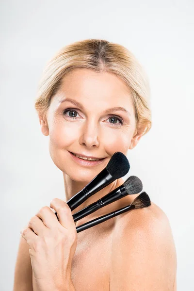 Beautiful and smiling mature woman looking at camera and holding cosmetic brushes on grey background — Stock Photo