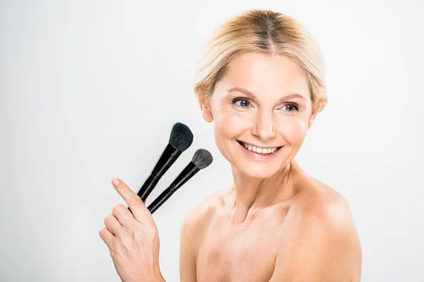 Hermosa y sonriente mujer madura mirando hacia otro lado y la celebración de cepillos cosméticos sobre fondo gris - foto de stock