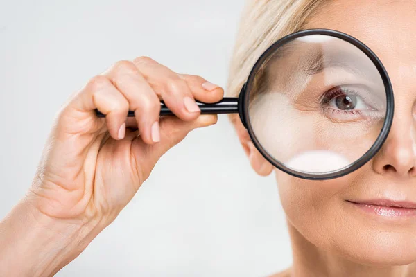 Selektiver Fokus der reifen Frau, die in die Kamera blickt und Lupe auf grauem Hintergrund hält — Stockfoto