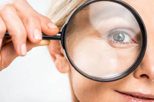 Selective focus of mature woman looking at camera and holding loupe isolated on grey — Stock Photo