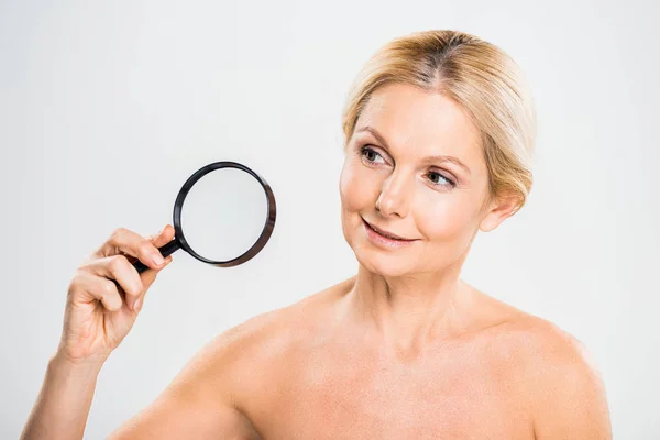 Beautiful and blonde mature woman holding loupe and looking away on grey background — Stock Photo