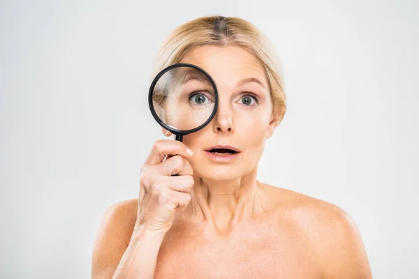 Beautiful and surprised mature woman holding loupe and looking at camera on grey background — Stock Photo