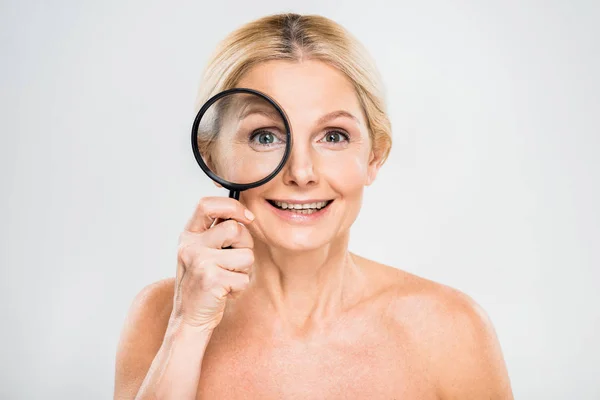 Beautiful and smiling mature woman holding loupe and looking at camera on grey background — Stock Photo