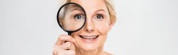 Panoramic shot of beautiful and smiling mature woman holding loupe and looking at camera on grey background — Stock Photo