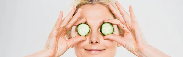 Panoramic shot of beautiful and blonde mature woman holding cut cucumber on grey background — Stock Photo