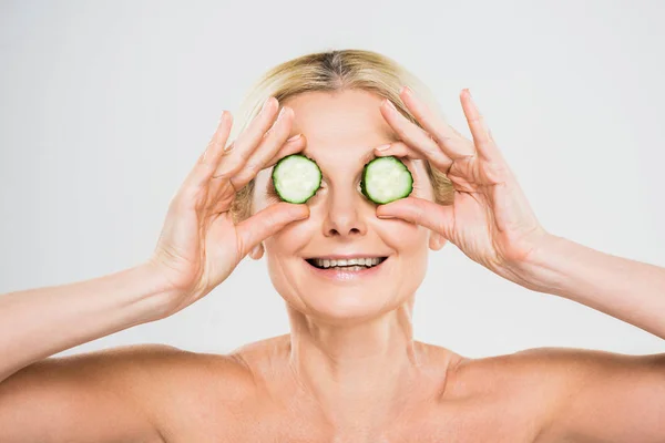 Hermosa y sonriente mujer madura sosteniendo pepino cortado sobre fondo gris - foto de stock