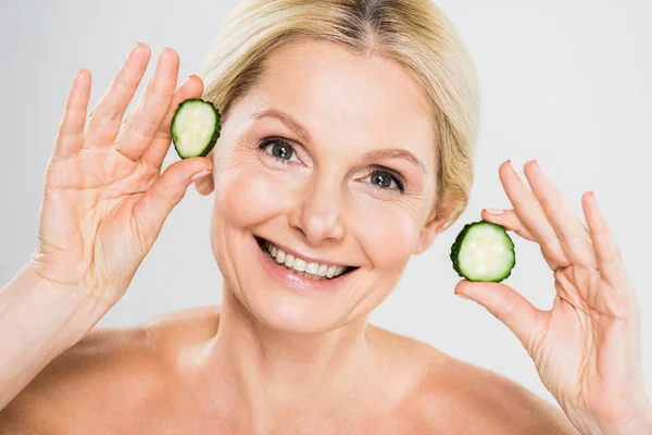 Beautiful and smiling mature woman holding cut cucumber and looking at camera on grey background — Stock Photo
