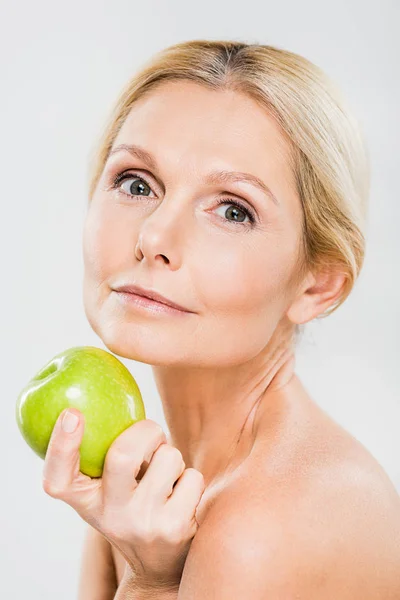 Красива і зріла жінка тримає зелене стигле яблуко і дивиться на камеру ізольовано на сірому — стокове фото