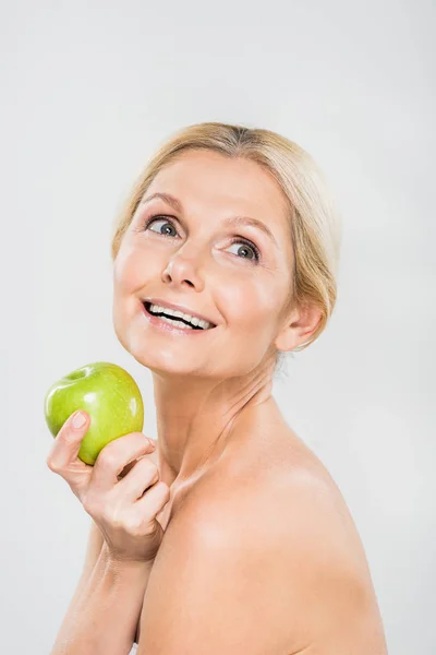 Beautiful and smiling mature woman holding green ripe apple and looking away isolated on grey — Stock Photo