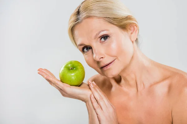 Beautiful and mature woman holding green apple and looking at camera isolated on grey — Stock Photo