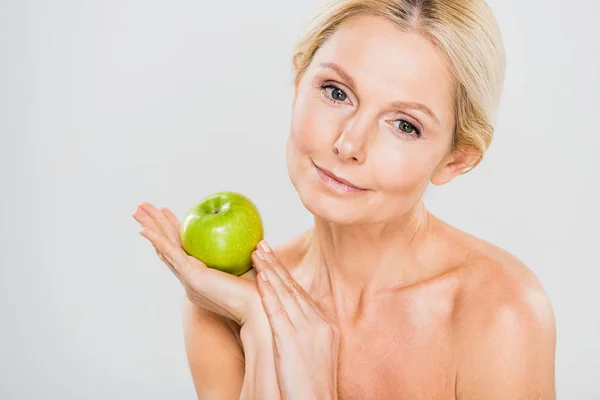 Beautiful and mature woman holding green ripe apple and looking away on grey background — Stock Photo