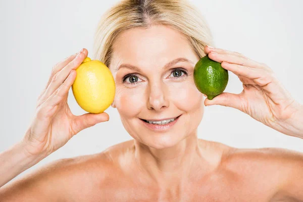 Hermosa y sonriente mujer madura sosteniendo verde lima madura y limón y mirando a la cámara sobre fondo gris - foto de stock