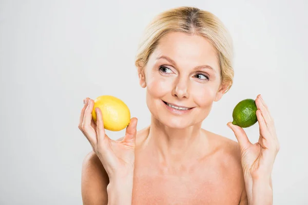 Hermosa y sonriente mujer madura sosteniendo lima madura y limón y mirando hacia otro lado sobre fondo gris - foto de stock