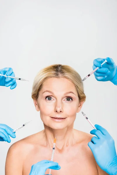 Hermosa y madura mujer mirando a la cámara con cosmetólogos sosteniendo jeringas - foto de stock