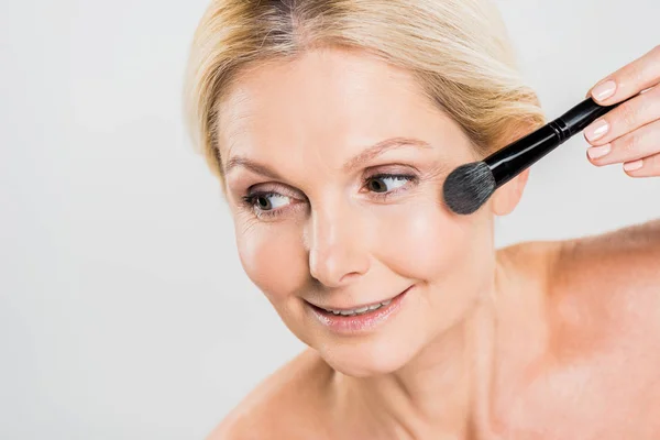 Beautiful and smiling woman applying blush with cosmetic brush and looking away isolated on grey — Stock Photo