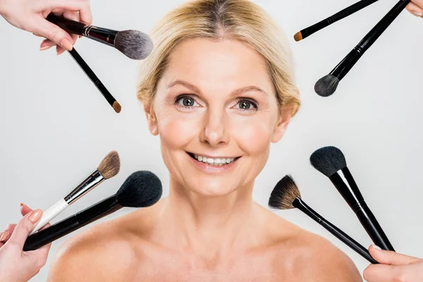 Hermosa y sonriente mujer mirando a la cámara y los artistas de maquillaje con cepillos cosméticos aislados en gris - foto de stock
