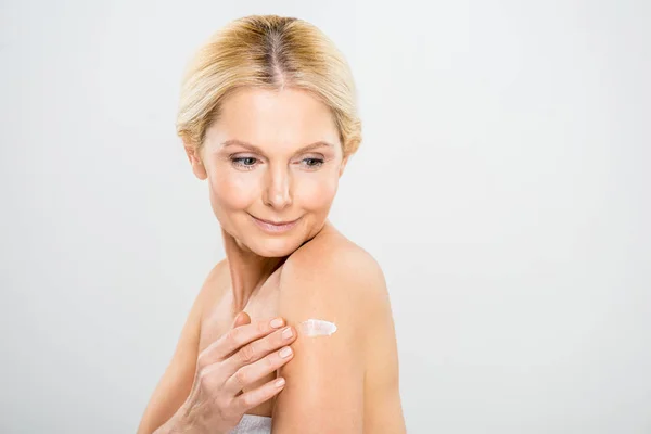Hermosa y madura mujer mirando hacia abajo y la aplicación de crema cosmética en el hombro - foto de stock